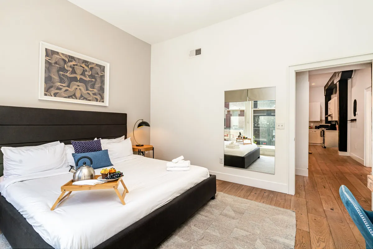 A stylish bedroom with a bed adorned with decorative pillows, a breakfast tray set for a leisurely morning, a large mirror, and modern wall art, bathed in soft natural light.
