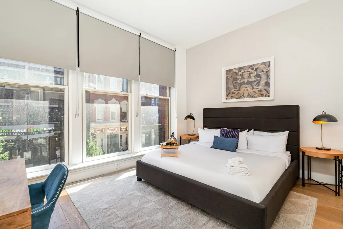 A serene, spacious bedroom with a plush bed dressed in white linens, large windows letting in natural light, tasteful artwork on the wall, and a dedicated desk and chair positioned nearby—ideal for work or study.