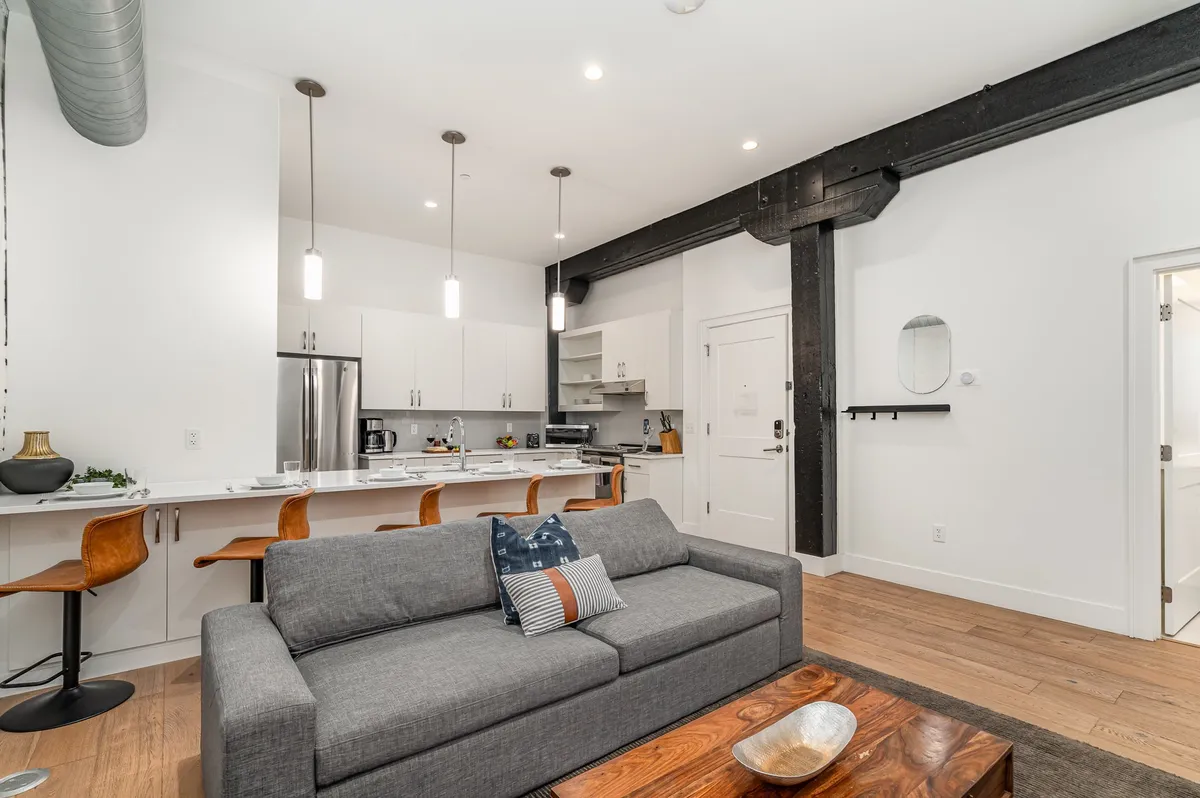 A cozy, modern living space featuring a plush sofa facing a wooden coffee table, bar stools at a kitchen island, and a sleek open kitchen. The setup suggests effortless transitions between cooking, entertaining, and relaxing, making it easy for a group to stay connected.