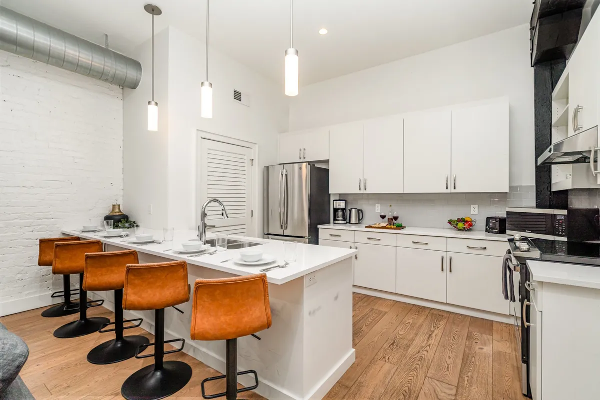 A bright, modern kitchen featuring an island with several bar stools, white cabinetry, and high-end appliances. The open counter space and inviting setup reinforce the idea of communal meals and gatherings.