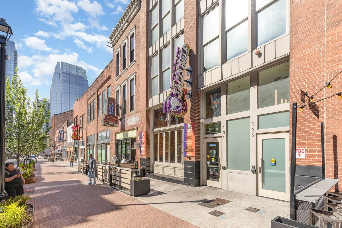 An energetic streetscape with restaurant facades, a Taco Bell sign, sidewalk seating, and street lamps bustling with people. The setting captures the lively Nashville atmosphere, perfect for strolling and exploring diverse eateries and local culture.