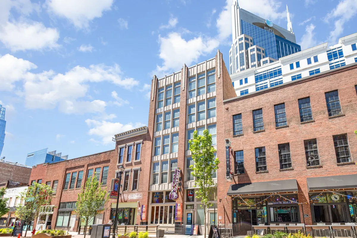 The image showcases a beautifully restored historic brick building nestled in the heart of downtown Nashville, with sleek modern skyscrapers nearby and lush street greenery that frames cozy outdoor seating areas. The lively street scene hints at nearby cafes, boutique shops, and easy walkability to the city's famous attractions.